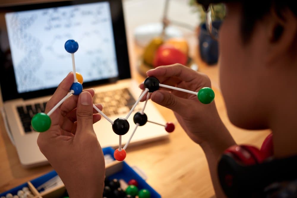 Student assembling a ball-and-stick molecular model on a desk with a laptop.