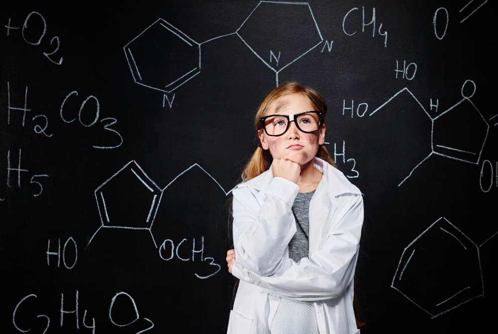 Young girl in a lab coat and glasses standing in front of a blackboard covered in chemistry formulas