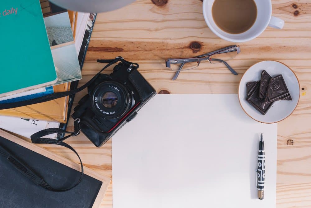 Overhead view of a wooden desk with a camera, books, coffee, glasses, chocolate, and a blank sheet of paper.